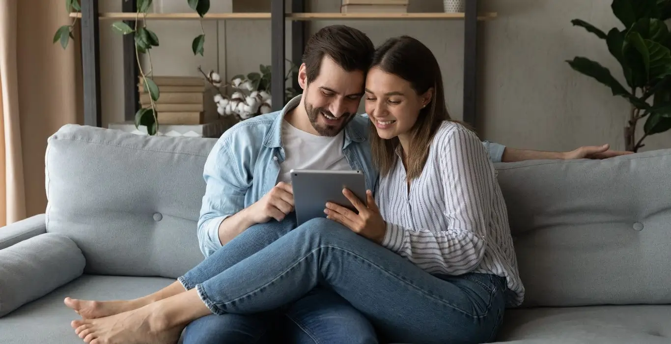 Jeune couple souriant consultant une tablette sur leur canapé pour imaginer leur future maison en Savoie.