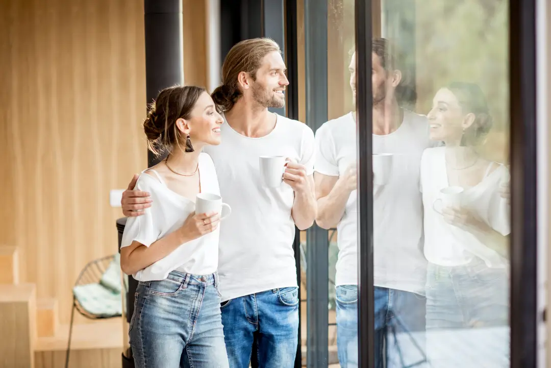 Jeune couple regardant par la fenêtre de leur maison moderne avec des mugs